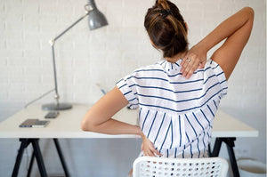 Woman sitting at a desk, holding her lower back and shoulder to show discomfort from poor posture.