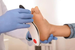 Podiatrist wearing blue gloves fitting a white orthotic insert with red heel padding under a patient’s bare foot during an assessment.