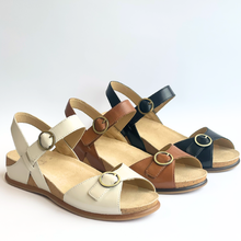 Three women's open-toe slingback wedge sandals arranged in a row on a light gray background. The sandals feature wide straps with round gold-tone buckle details, cork-like midsoles, and tan footbeds.